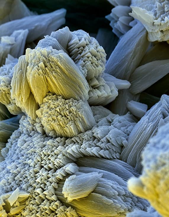 A false-colour electron microscope image of limescale: interlocking calcium carbonate crystals in pale blue-grey and yellow, stacked in blocky plates like the splayed pages of books, with jagged stepped edges.