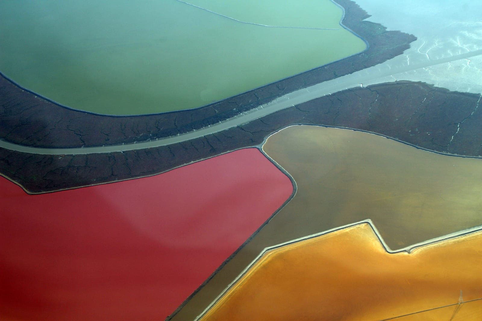 An aerial photograph of salt evaporation ponds forming irregular blocks of vivid colour separated by narrow earth levees — one pond deep crimson, another olive green, others tan and burnt orange, with a pale road curving between them.